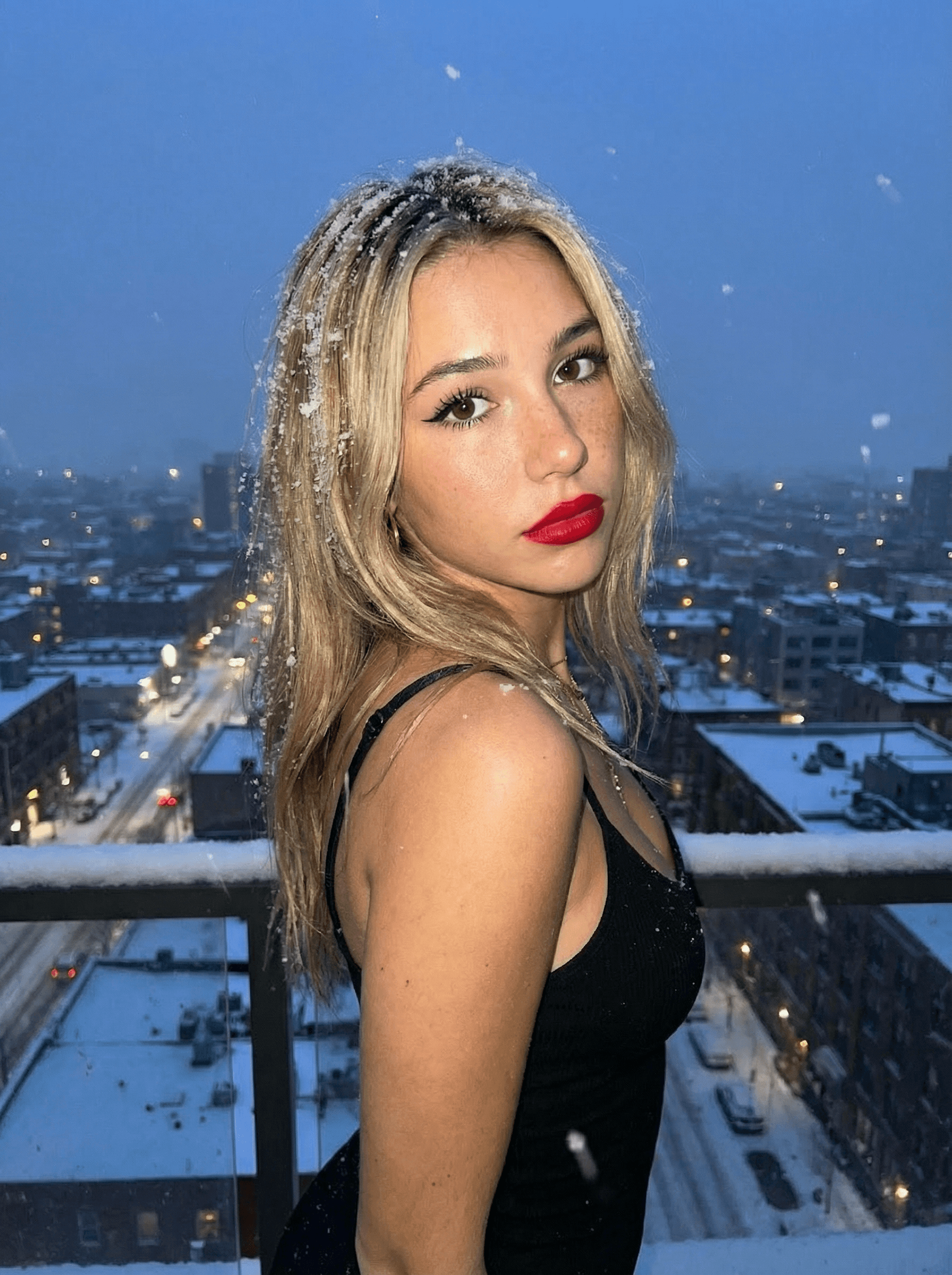create a great photo of my influencer @Sarah and she is in a sundress on top of a rooftop with the city overlooking the view and its snowing in the background. close up shot she is looking at us with puppy eyes. There is slight snow fall in her hair, she has slight straps on her bra. She has red lipstick on visible and mascara and eyeliner wings on. Flash on the camera, exposure