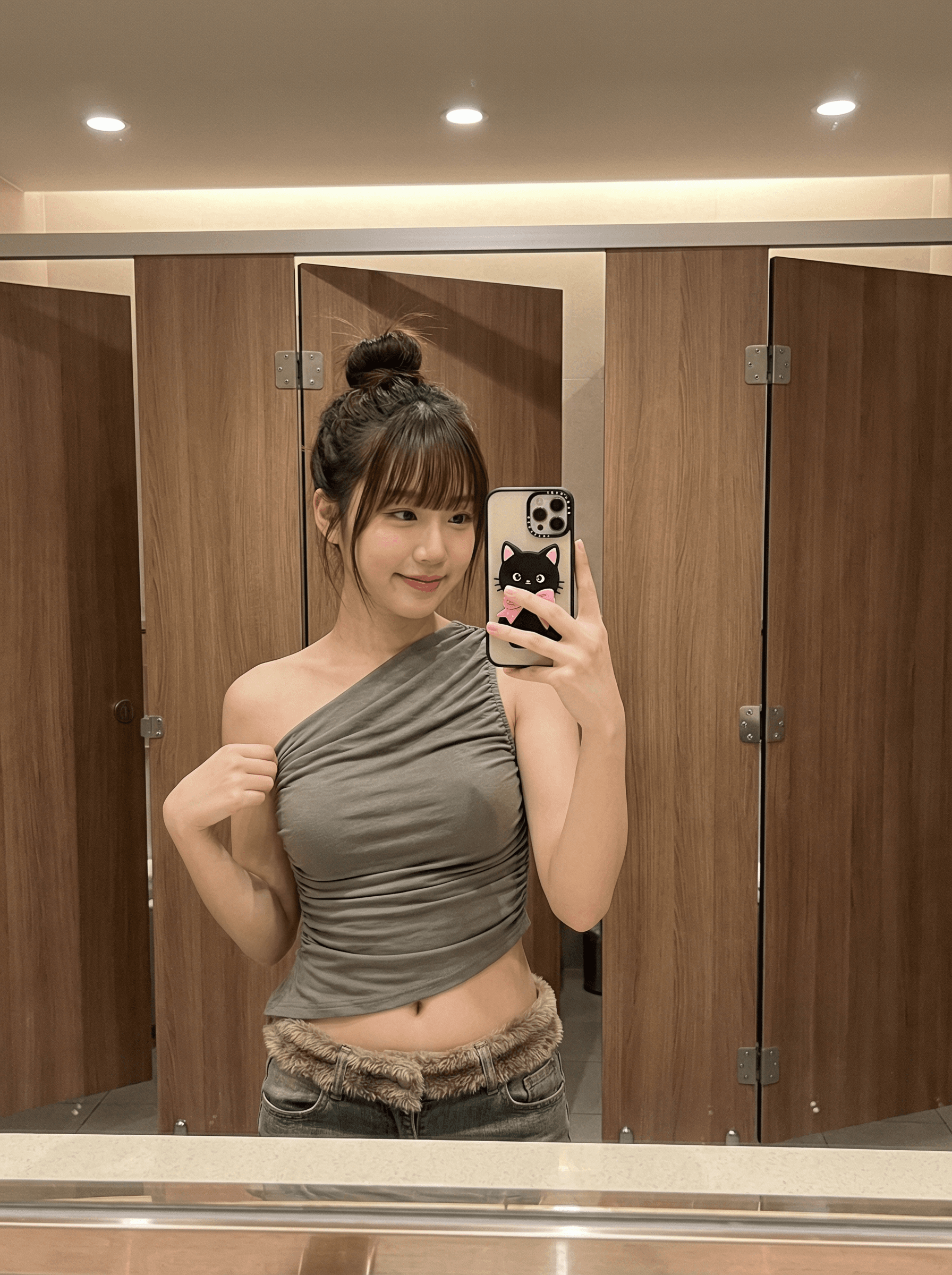 {
"scene": {
"description": "Stylish restroom dressing area captured via mirror.",
"environment": "Warm wood-laminate stall doors and soft countertop reflections.",
"mood": "Shy-playful confidence; cozy 'Gamseong' (감성) aesthetic."
},
"aesthetic": {
"style": "Authentic smartphone mirror selfie.",
"look": "Warm interior color, gentle roll-off; minimal grain; filmic soft contrast."
},
"lighting": {
"description": "Ceiling LEDs providing warm ambience; add subtle front fill from mirror to open eye sockets; maintain realistic specular highlights."
},
"subject": {
"demographics": {
"ethnicity": "East Asian (Korean)",
"age": "adult",
"build": "Very slender, petite with defined waist."
},
"appearance": {
"hair": "Messy top-bun with full fringe; a few loose side strands.",
"skin": "Natural, dewy finish with preserved skin texture."
},
"pose": {
"type": "Mirror selfie, standing.",
"action": "Gentle smile with lips slightly pressed; left hand lightly lifting ruched hem; right hand holding phone.",
"framing": "Waist-up 3:4 vertical; slight downward camera angle; midriff visible."
},
"wardrobe": {
"top": "Asymmetrical olive-gray ruched one-shoulder top.",
"bottom": "Low-rise jeans with faux-fur waistband trim."
},
"accessories": {
"jewelry": "Thin bracelets, rings, and delicate hoop earrings.",
"phone": "Smartphone with black cat case featuring a pink bow (Hello-Kitty style)."
}
},
"props_in_scene": {
"background": "Wood-grain stall doors with verticals kept true; metal hinges subtly visible.",
"counter": "Keep any edge reflections soft and realistic."
},
"camera_technical": {
"requirements": "Correct mirror reversal; maintain natural body proportions; no duplicated phones/hands.",
"capture": "Smartphone wide lens ~24–26mm equiv, f/1.8 look, ISO ~250, 1/125s, WB 4200–4400K.",
"composition": "Subject centered with slight lean; keep fur texture readable; ensure phone and hand pose feel natural.",
"retouching": "Clean minor blemishes only; preserve fabric micro-wrinkles and fur fibers; avoid haloing.",
"avoid": "Warped doors, heavy vignettes, oversharpening, cartoon smoothing, text/watermarks."
}
}