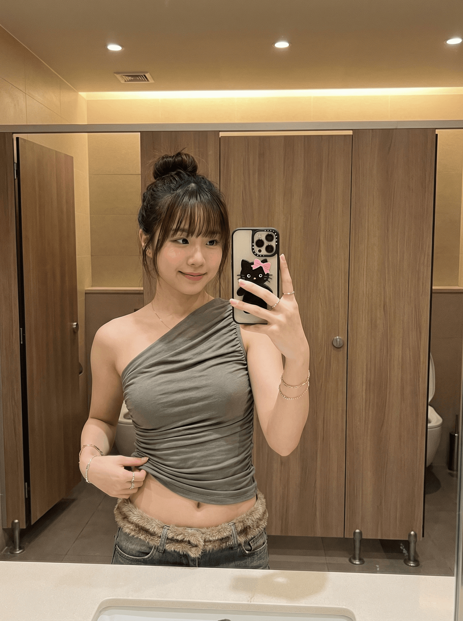 {
"scene": {
"description": "Stylish restroom dressing area captured via mirror.",
"environment": "Warm wood-laminate stall doors and soft countertop reflections.",
"mood": "Shy-playful confidence; cozy 'Gamseong' (감성) aesthetic."
},
"aesthetic": {
"style": "Authentic smartphone mirror selfie.",
"look": "Warm interior color, gentle roll-off; minimal grain; filmic soft contrast."
},
"lighting": {
"description": "Ceiling LEDs providing warm ambience; add subtle front fill from mirror to open eye sockets; maintain realistic specular highlights."
},
"subject": {
"demographics": {
"ethnicity": "East Asian (Korean)",
"age": "adult",
"build": "Very slender, petite with defined waist."
},
"appearance": {
"hair": "Messy top-bun with full fringe; a few loose side strands.",
"skin": "Natural, dewy finish with preserved skin texture."
},
"pose": {
"type": "Mirror selfie, standing.",
"action": "Gentle smile with lips slightly pressed; left hand lightly lifting ruched hem; right hand holding phone.",
"framing": "Waist-up 3:4 vertical; slight downward camera angle; midriff visible."
},
"wardrobe": {
"top": "Asymmetrical olive-gray ruched one-shoulder top.",
"bottom": "Low-rise jeans with faux-fur waistband trim."
},
"accessories": {
"jewelry": "Thin bracelets, rings, and delicate hoop earrings.",
"phone": "Smartphone with black cat case featuring a pink bow (Hello-Kitty style)."
}
},
"props_in_scene": {
"background": "Wood-grain stall doors with verticals kept true; metal hinges subtly visible.",
"counter": "Keep any edge reflections soft and realistic."
},
"camera_technical": {
"requirements": "Correct mirror reversal; maintain natural body proportions; no duplicated phones/hands.",
"capture": "Smartphone wide lens ~24–26mm equiv, f/1.8 look, ISO ~250, 1/125s, WB 4200–4400K.",
"composition": "Subject centered with slight lean; keep fur texture readable; ensure phone and hand pose feel natural.",
"retouching": "Clean minor blemishes only; preserve fabric micro-wrinkles and fur fibers; avoid haloing.",
"avoid": "Warped doors, heavy vignettes, oversharpening, cartoon smoothing, text/watermarks."
}
}